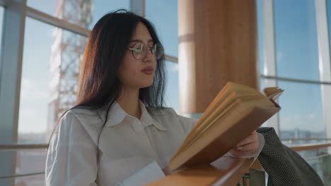 high school student wearing glasses reading open book while leaning on wooden rail in sunlit indoor setting, with blurred view of modern urban skyline visible through large glass windows behind her