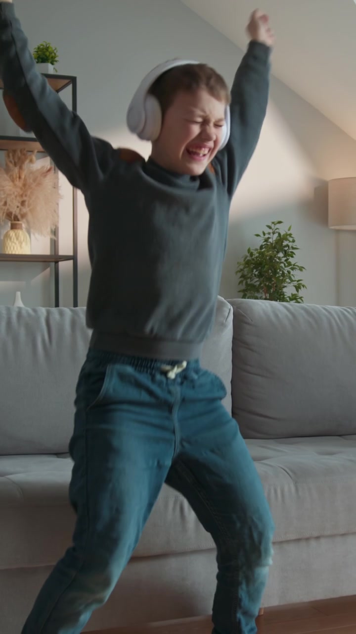 Young boy wearing headphones enthusiastically dances while looking at his phone screen in a living
