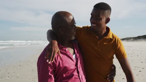 African-american-senior-father-and-teenage-son-standing-on-beach-embracing-and-talking