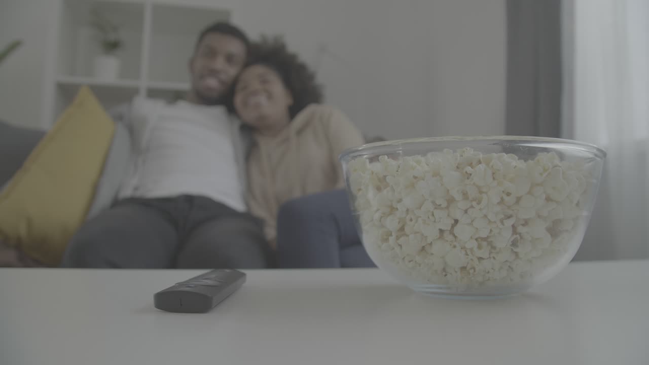 Details of a remote control a bowl of popcorn. In the background, a young african american couple enjoy while watching a movie