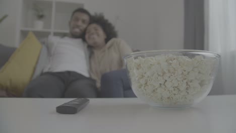 details of a remote control a bowl of popcorn. in the background, a young african american couple enjoy while watching a movie