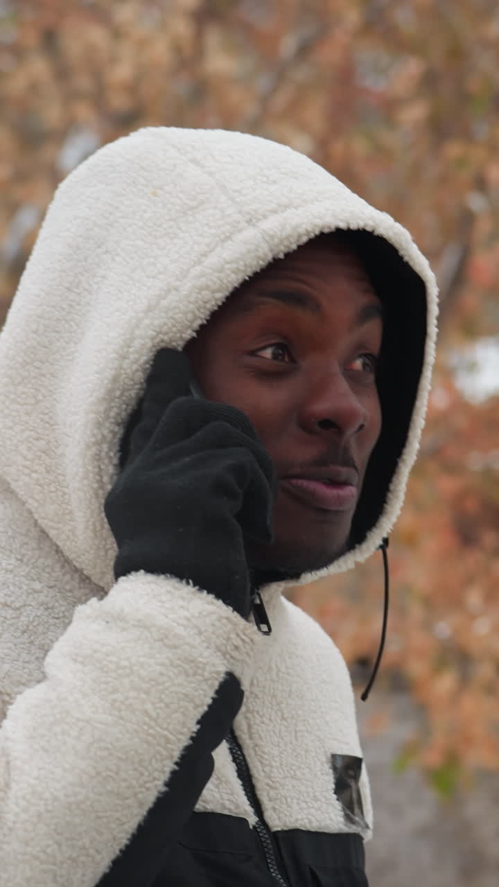 vista lateral de un joven con capucha blanca conversando por teléfono con alguien, sonriendo y comprometido, fondo borroso con árboles y edificios