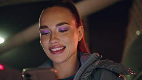 modelo positivo sonriendo cámara sosteniendo teléfono móvil en la noche oscura al aire libre.