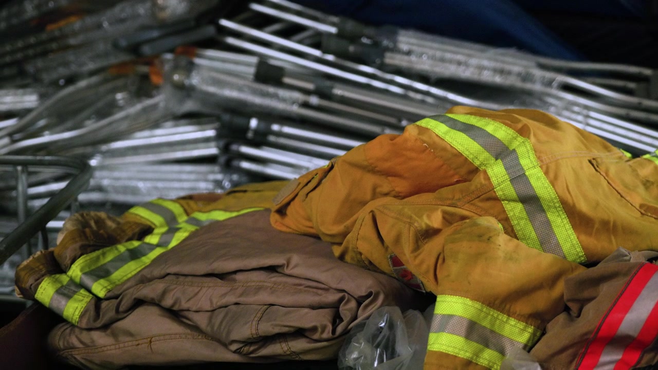 Una colección de chaquetas y equipo de bombero yace sobre herramientas de metal en un espacio de almacenamiento. Las chaquetas muestran signos de uso y están dispuestas de forma casual.