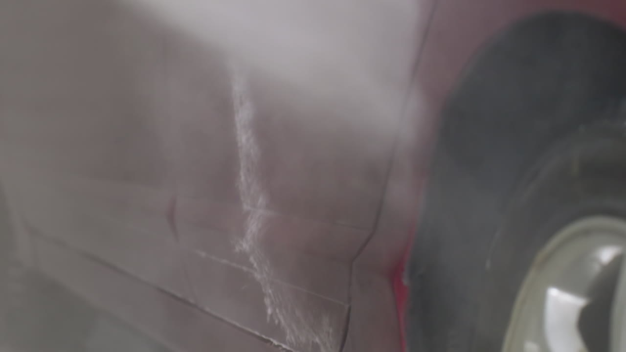 Macro close up of car being rinsed with high pressure water on side door revealing dynamic liquid flow and suds dripping on glossy red surface inside garage during professional cleaning process