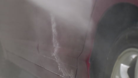macro close up of car being rinsed with high pressure water on side door revealing dynamic liquid flow and suds dripping on glossy red surface inside garage during professional cleaning process