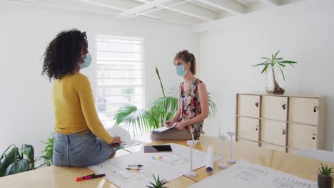 Two-woman-wearing-face-mask-discussing-over-documents-on-table-at-office