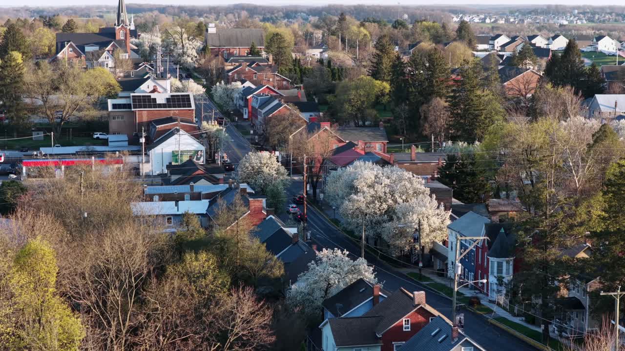 Luchtopname met drone van een rustige Amerikaanse wijk in Pennsylvania, met charmante huizen, bloeiende lentebomen en zacht ochtendzonlicht. Vredige voorstedelijke sfeer. Breedbeeldopname. Rustige, serene scène.