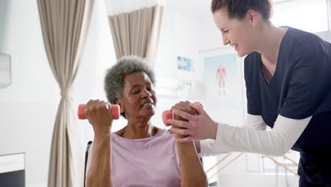 Diverse-senior-woman-exercising-with-weights-and-female-doctor-advising-in-hospital,-slow-motion