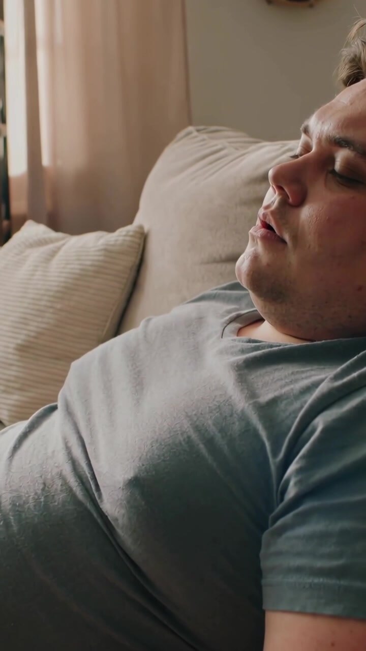 Vertical video: Streaming daylight lulling male dozing on sofa at home in teal tee with cushion