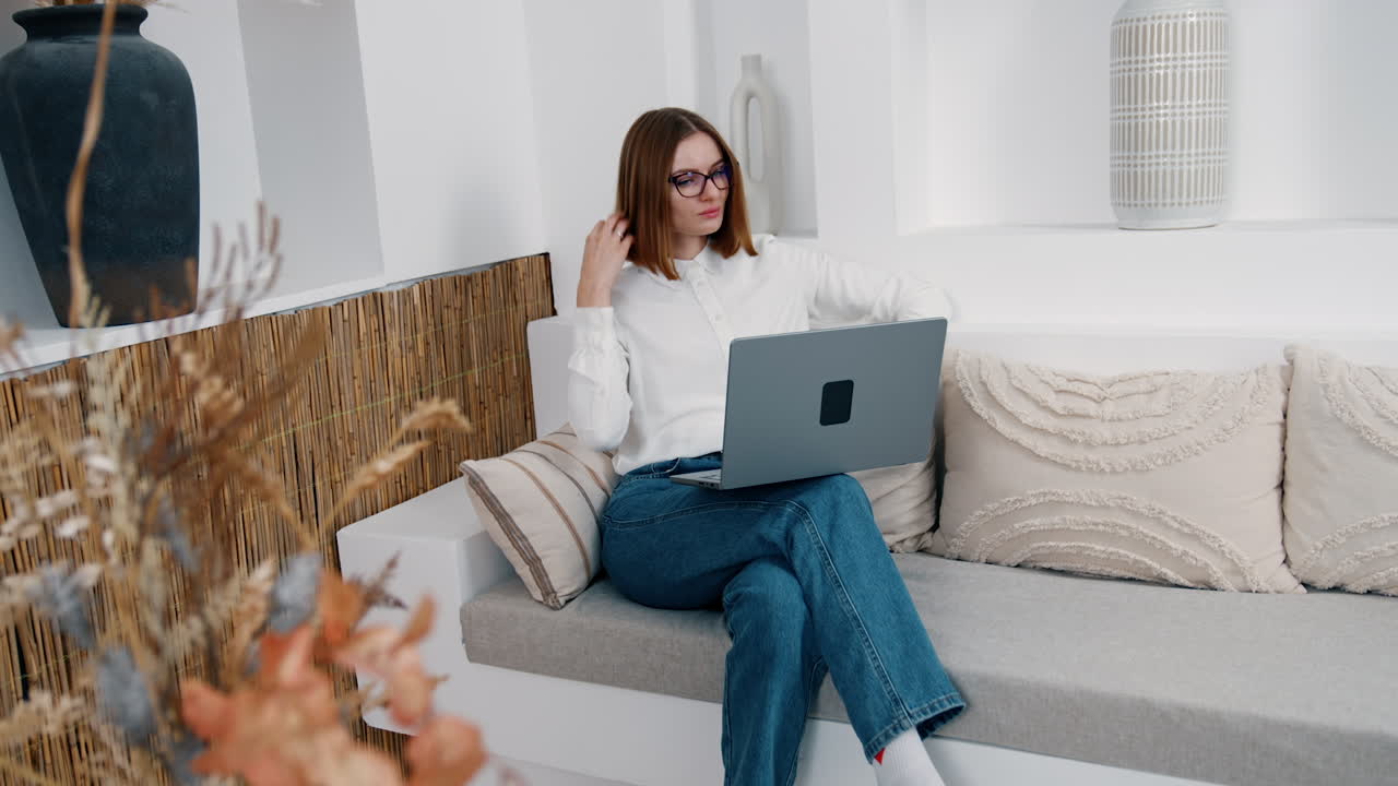 Caucasian lady sits comfortably on the sofa and touches her hair preparing for the video call. Remote working conditions at home.