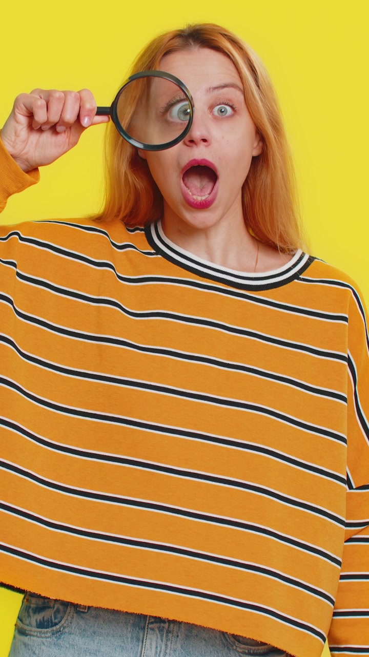 Young adult woman holds magnifying glass near face looking at camera with big zoomed eye analyzing