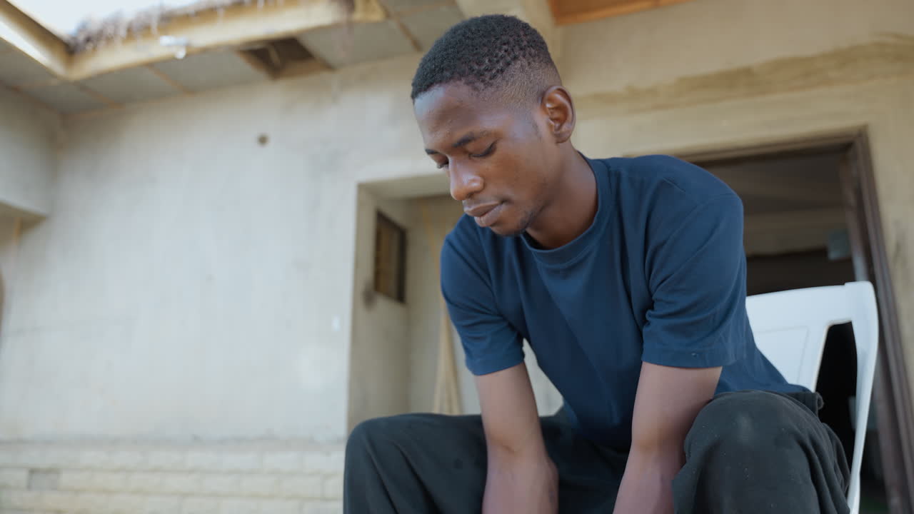 Wide shot black man after chores, seated on house steps, concrete stoop and unfinished plaster exterior, navy shirt, downward gaze, hands resting on knees, midday light, quiet pause between tasks
