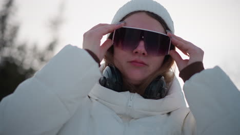 close up side view of young woman wearing tinted sunglasses and headphones around neck, as she moves to music under bright winter sky wind tousles hair rosy cheeks and manicured nails glisten