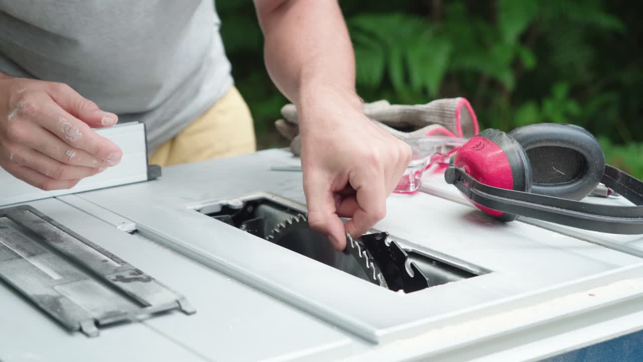 Hands adjusting circular saw blade on outdoor table saw, safety gear gloves, glasses, ear protection nearby, carpentry preparation task
