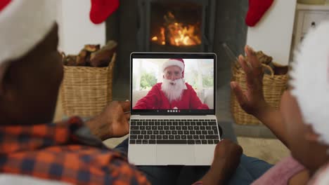 Afroamerikanisches-Paar-Mit-Weihnachtsmützen-Nutzt-Laptop-Für-Einen-Weihnachtsvideoanruf-Mit-Dem-Weihnachtsmann-Auf-Dem-Bildschirm