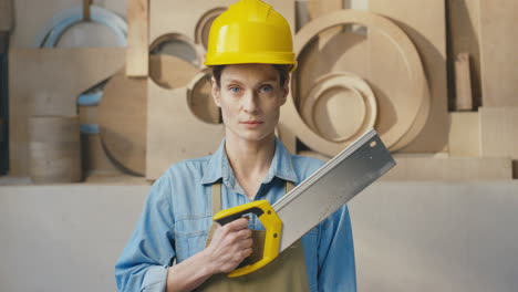 portrait of caucasian woman in helmet holding saw and looking at camera in carpentry workshop