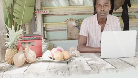 Happy-african-american-man-using-laptop-behind-the-counter-of-surf-rental-beach-shack,-slow-motion