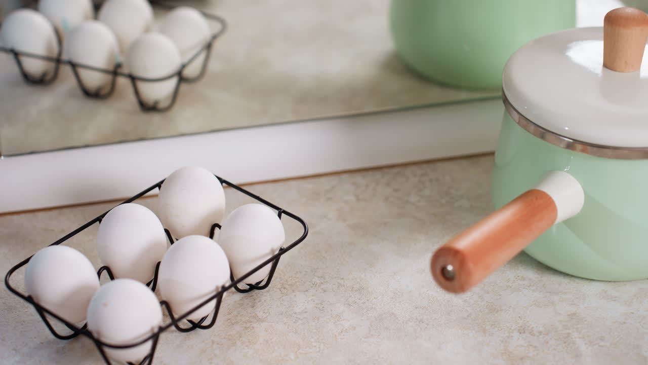 Brightly arranged kitchen counter with six white eggs in black wire basket next to mint green cooking pot with wooden handle, reflected clearly in large mirror
