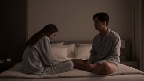 a couple in pajamas sits on a bed in a dimly lit room, appearing to have a serious discussion