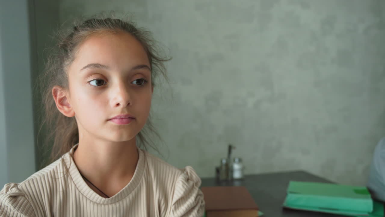 Focused girl raises hand while sitting at table with boy in background, green folders and book on table, modern room setting, suggesting classroom or homework scene, concentration, and learning effort