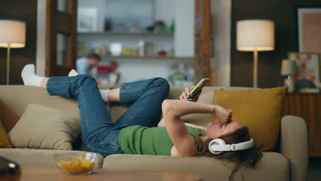 Lazy girl enjoying headset music laying couch. Carefree lady browsing smartphone