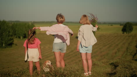 back view of family standing on hilly farmland under clear sky as mother points something interesting in distance to her daughter, while another woman stands in background holding flowers