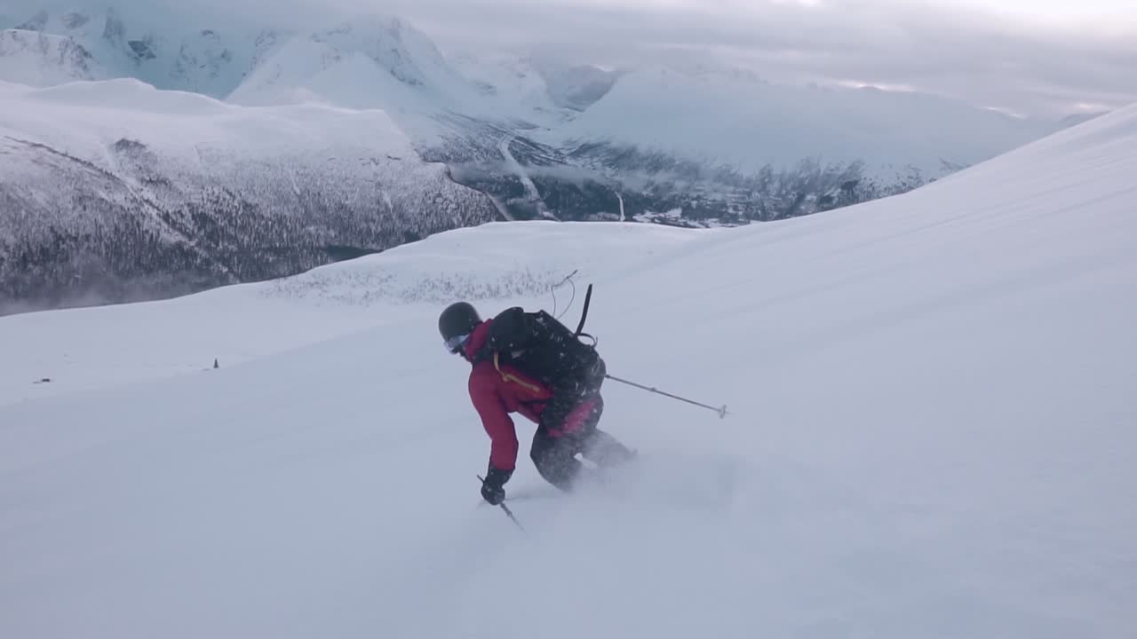 Ripresa al rallentatore di un uomo con giacca rossa che scia in discesa, circondato da montagne innevate e uno sfondo semi soleggiato coperto di nuvole-2