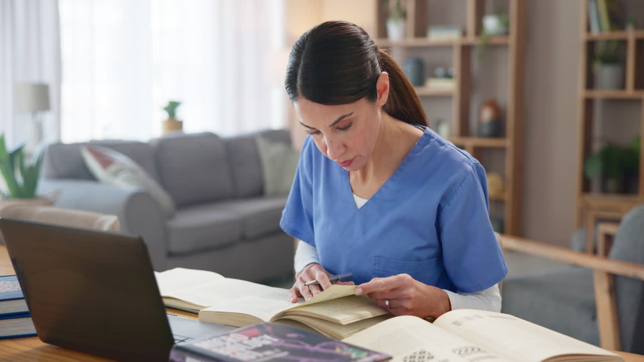 una mujer con uniforme de enfermera se sienta en un escritorio en su sala de estar, estudiando de libros y una computadora portátil.