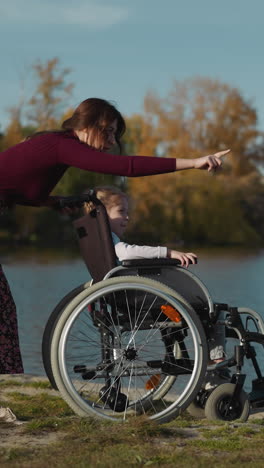 focused mother speaks to sitting in wheelchair little daughter. woman points finger and shows fishing gesture entertaining preschooler side view