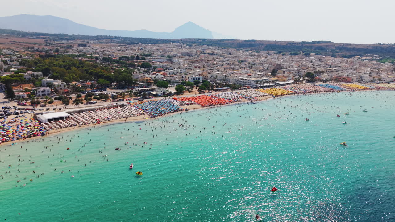 Aerial view of a crowded beach with colorful umbrellas near a town and mountains