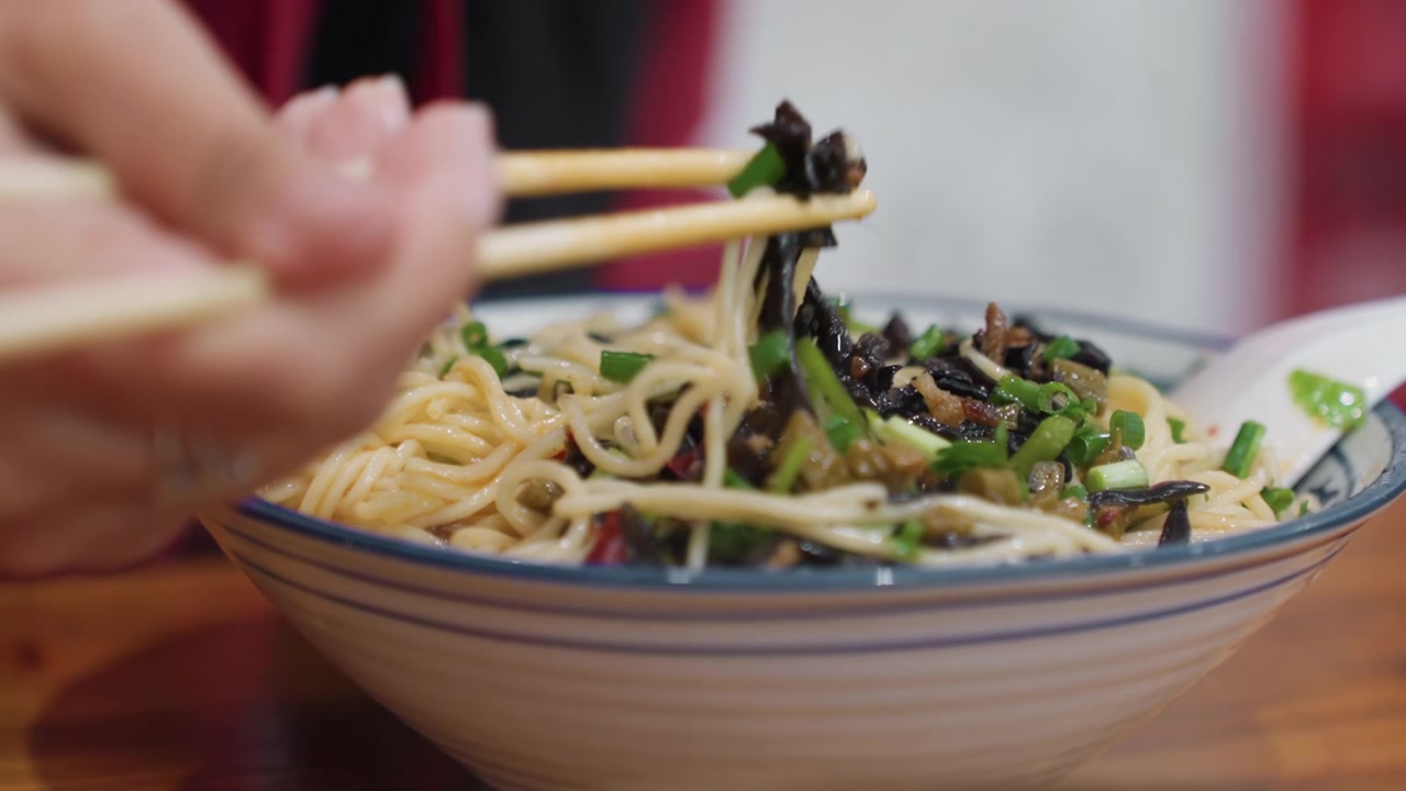 Primo piano di ciotola, noodles, bacchette, mani che sollevano fili di ramen, guarnizione di cipollina e alghe su un caldo tavolo di legno, brodo saporito che luccica nella ciotola di ceramica, movimento lento e intimo del mangiare, ristorante informale