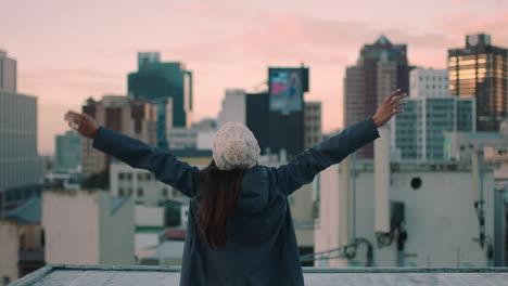 happy young woman on rooftop with arms raised celebrating freedom enjoying independent lifestyle looking at beautiful view of city skyline at sunset