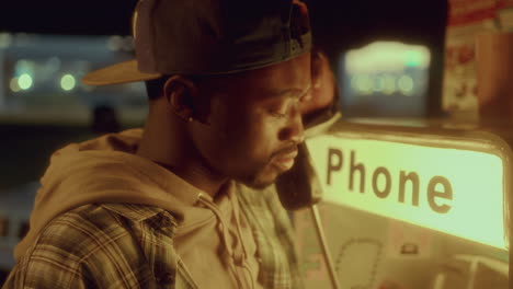 hombre negro hablando en un teléfono público iluminado en la calle bajo un paso elevado débilmente iluminado