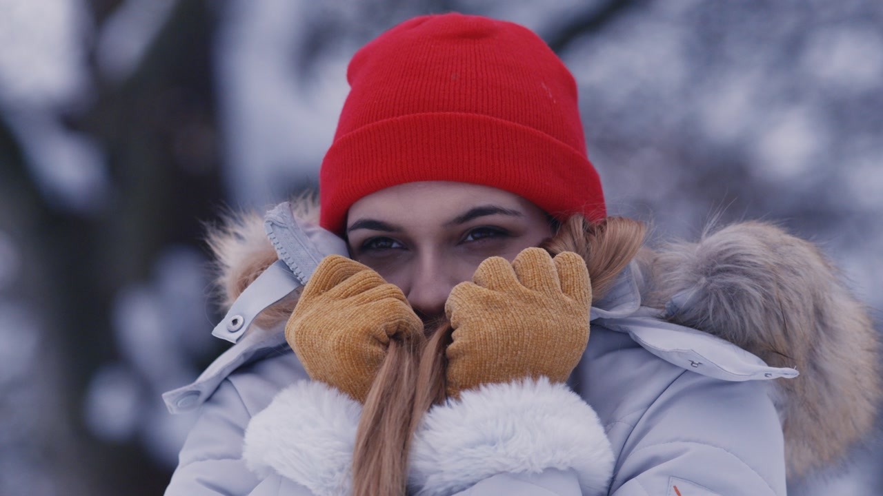 A woman in a red beanie and fur-trimmed jacket pulls her collar up to hide from the cold in a snowy forest.
