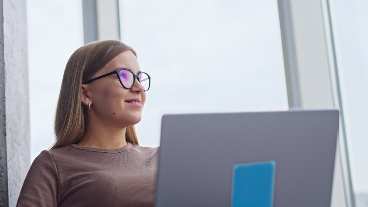 Confident relaxed blonde woman working on computer. Female looks into window for a moment and continues her work smiling. Low angle view.