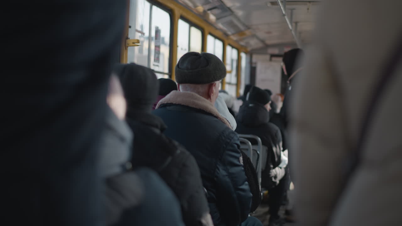 Winter commute in moving bus shows people in heavy coats seated and standing, urban interior with window light, bundled passengers traveling through city during cold ride, soft perspective