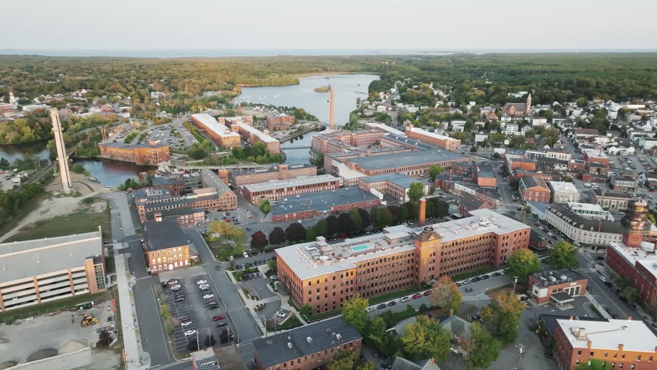 Aerial view of downtown Biddeford, Maine featuring historic mill buildings, the Saco River, and a vibrant small cityscape surrounded by forest and coastline.