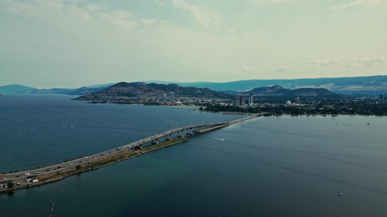 High Aerial over Okanagan Lake entire view William R Bennett Bridge Central Okanagan BC calm blue water traffic cars trucks commuting mountainous background light blue sky wispy clouds