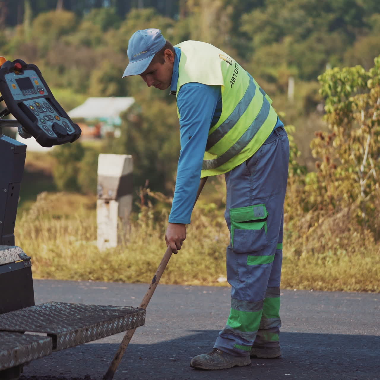 Close-up part of asphalt paver and a worker. Worker man in protective clothing works with special instrument after paver machine on the road.