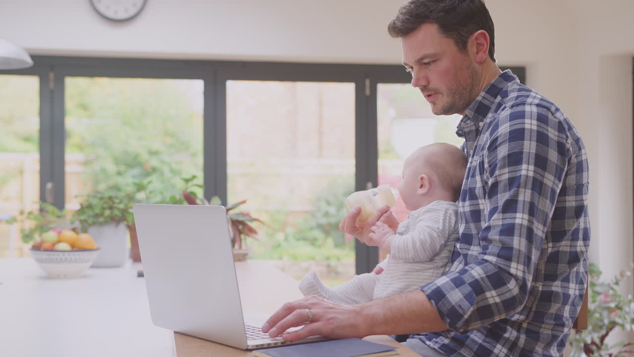 Working father using laptop at home whilst feeding baby son sitting on ...