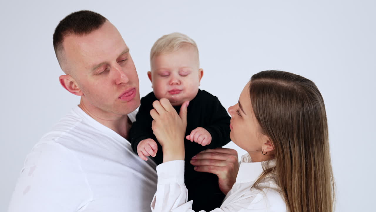 Funny Caucasian parents making faces copying their cute infant baby. Family portrait together at white backdrop.
