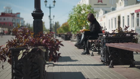 white girl seated in public space flipping through the page of book thoughtfully, with background showing flower pot swaying gently in the wind, and pedestrians in the background