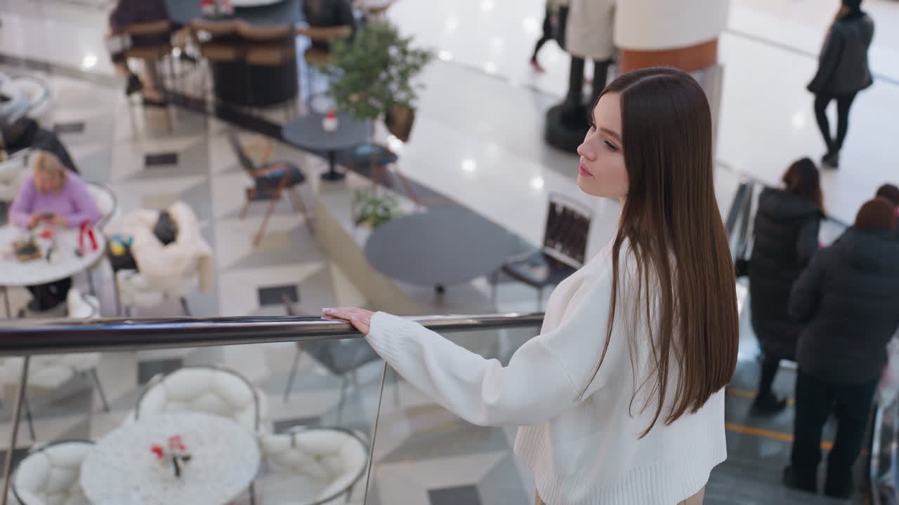 Stylish woman in white sweater descends escalator, gazing towards upscale restaurant on ground floor, elegant shopping mall ambiance with soft lighting, busy patrons, and modern interior