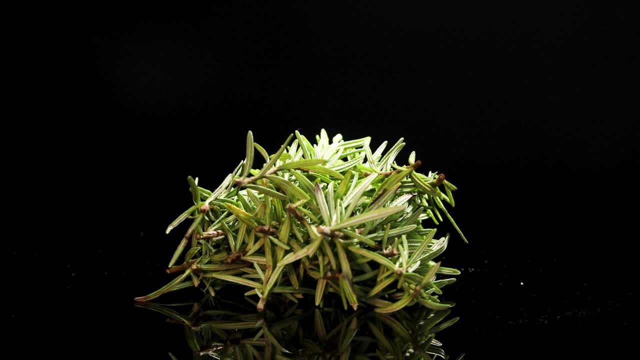 A bunch of rosemary rotates on a reflective black surface, highlighting its texture and vibrant green color
