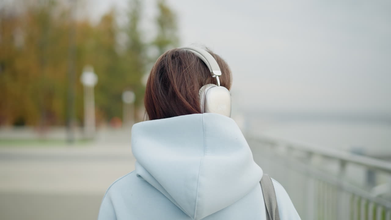 Real view of female artist in hoodie and headphones strolling, blurred withered tree and stainless rail in background, peaceful outdoor setting, casual walk, urban environment, relaxed mood