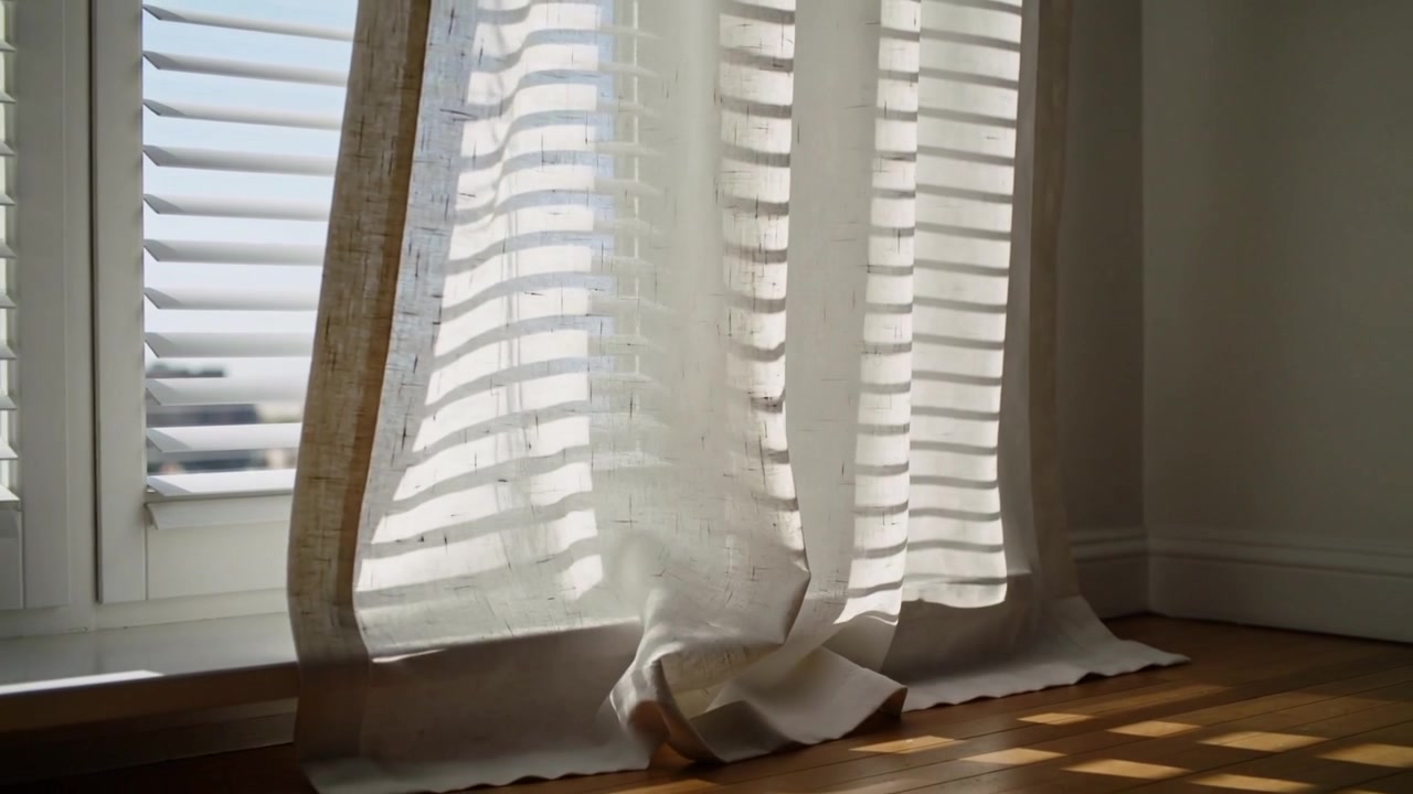 Cortina transparente con ribete beige meciéndose con la corriente de la ventana, levantando el dobladillo sobre el suelo de madera, con persianas proyectando sombras.