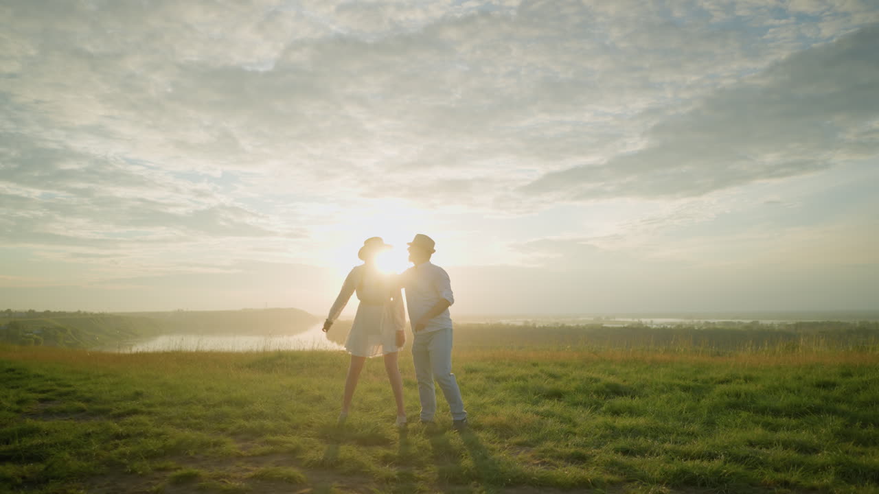 한 커플이 평온한 호수 근처의 잔디 언덕에서 한 에 춤을 추고, 황금빛을 즐기면서 안고 있습니다.  셔츠와 모자를 입은 남자, 흐르는  드레스를 입은 여자.
