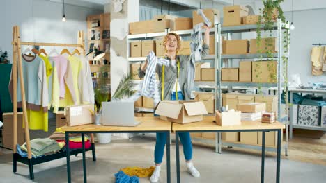 Young-happy-positive-caucasian-woman-clothing-designer-dancing-and-throwing-clothes-standing-in-own-shop-warehouse-with-boxes