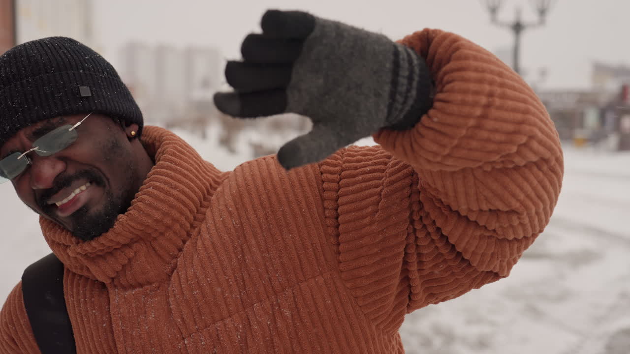 Uomo di colore cammina in una strada innevata, indossa un maglione marrone spesso, un berretto nero, occhiali da sole a specchio e guanti di lana, porta uno zaino mentre si ripara gli occhi dai fiocchi che cadono, vetrine e lampione.